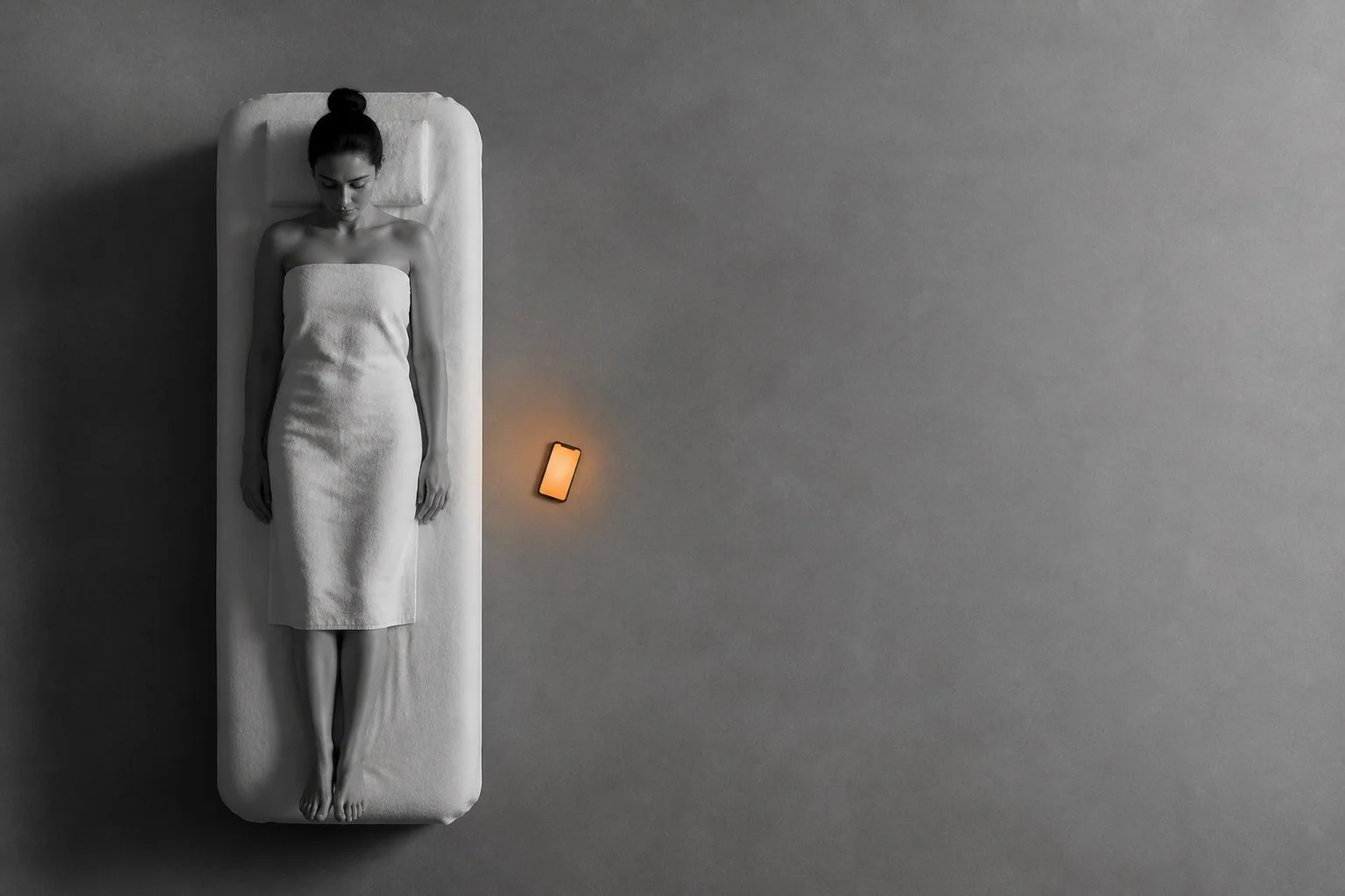 Black and white overhead view: someone resting on a treatment bed and, on the floor beside them, a phone lit up.